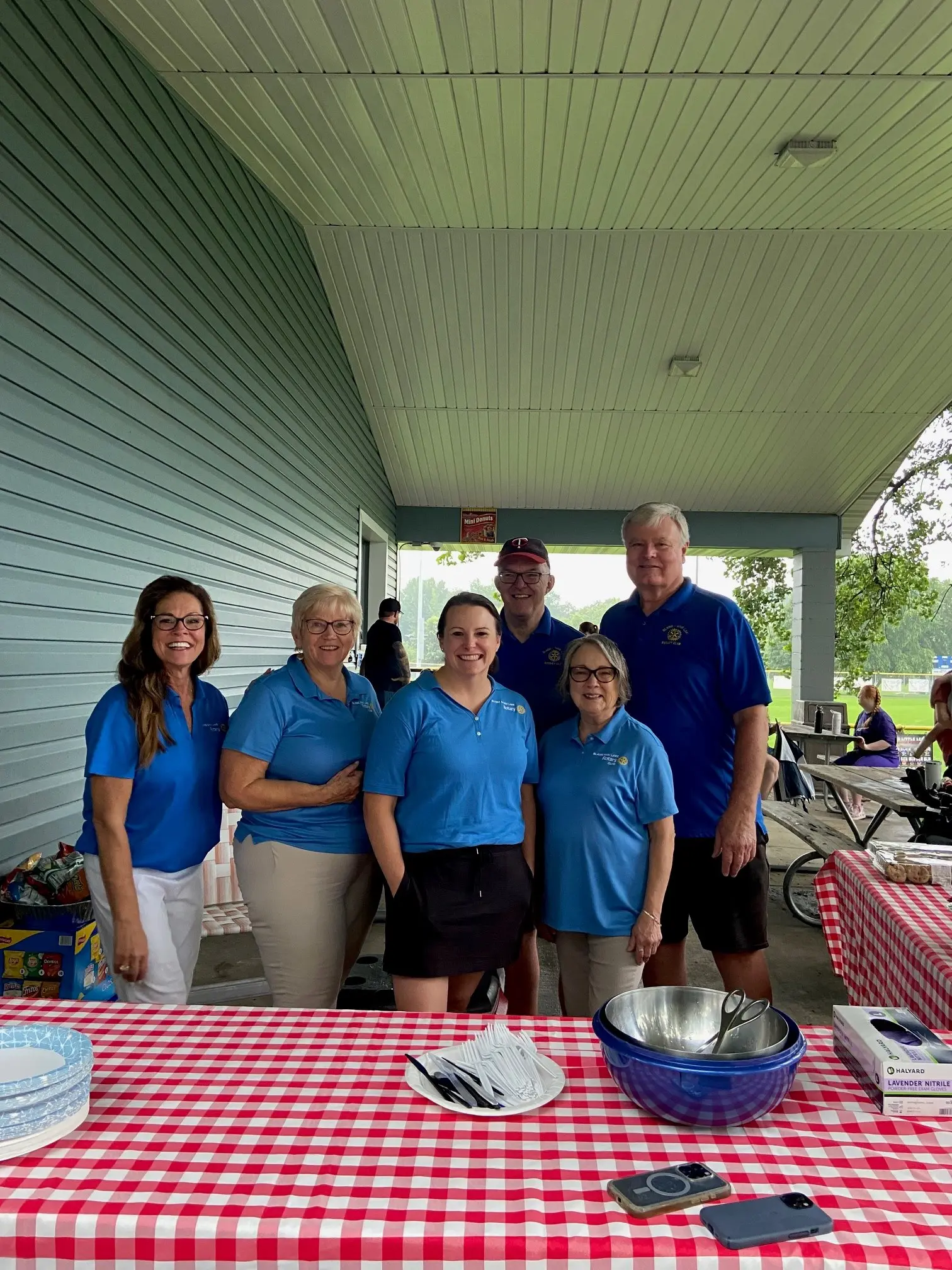 Six Rotarians pose behind a red and white checkered tablecloth while serving lunch to kids in the Challenger League (previously Miracle League)
